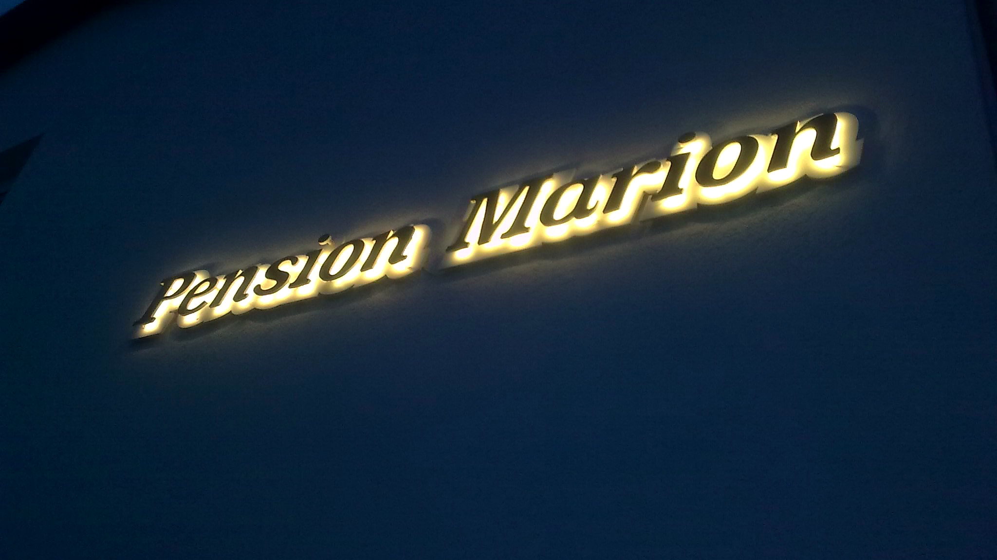 Schriftzug Pension Marion aus hinterleuchteten Einzelbuchstaben, warmweiss leuchtend an einer hellen Fassade zur blauen Stunde