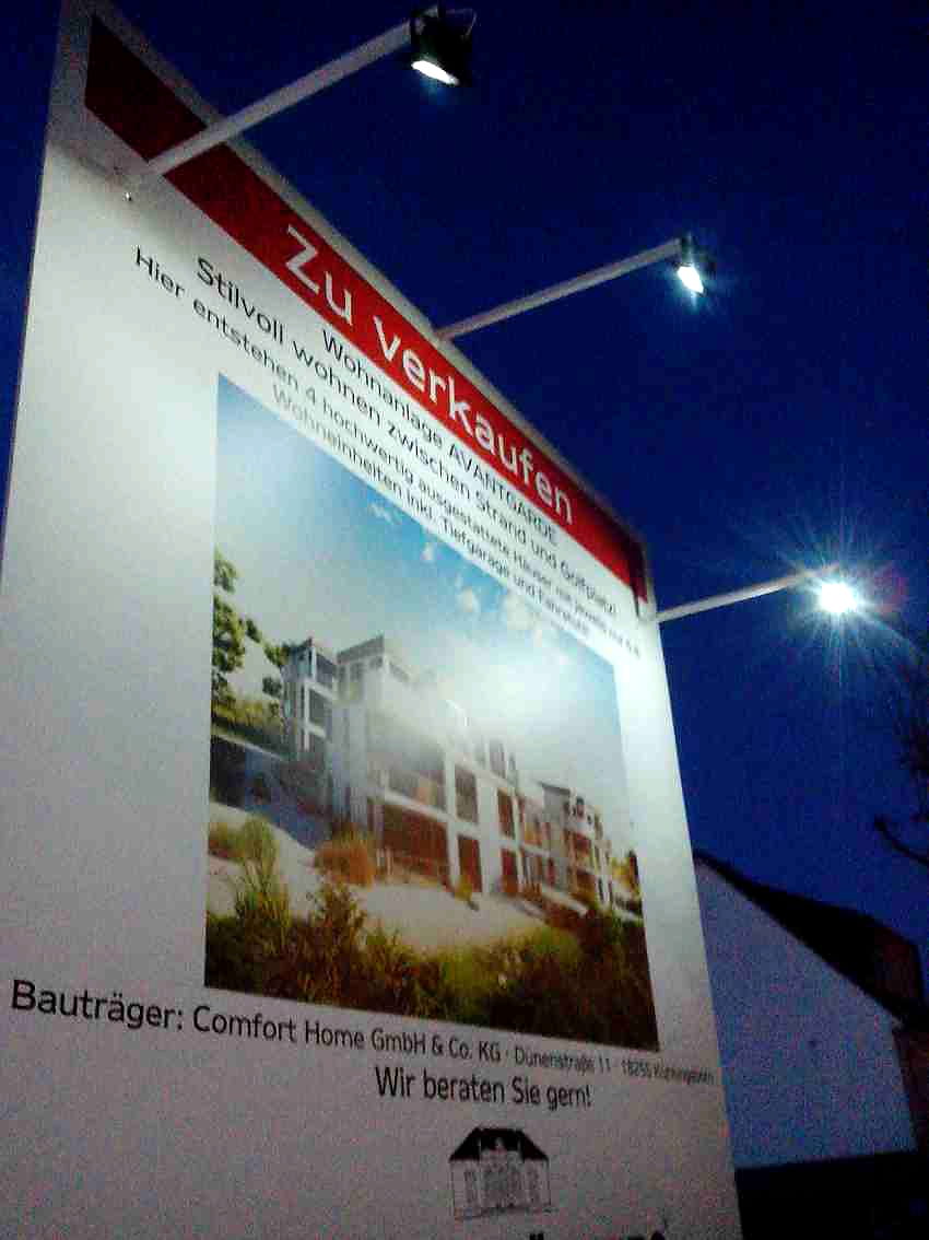 Beleuchtetes Bauschild der Wohnanlage Avantgarde in Binz mit Visualisierung und Strahlern zur blauen Stunde